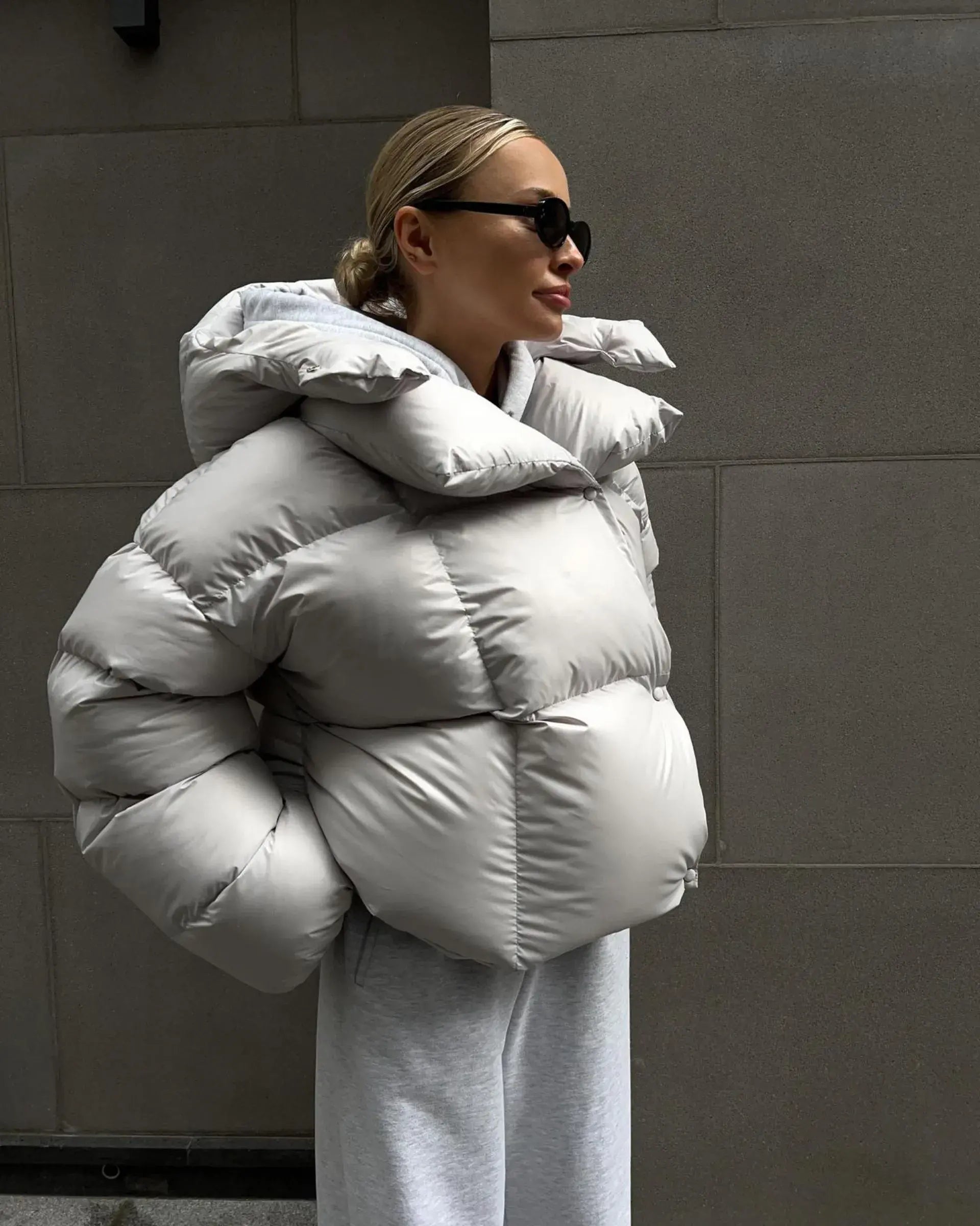 A woman wearing a hooded, puffer jacket in gray color with a high collar, sunglasses, and a casual outfit.